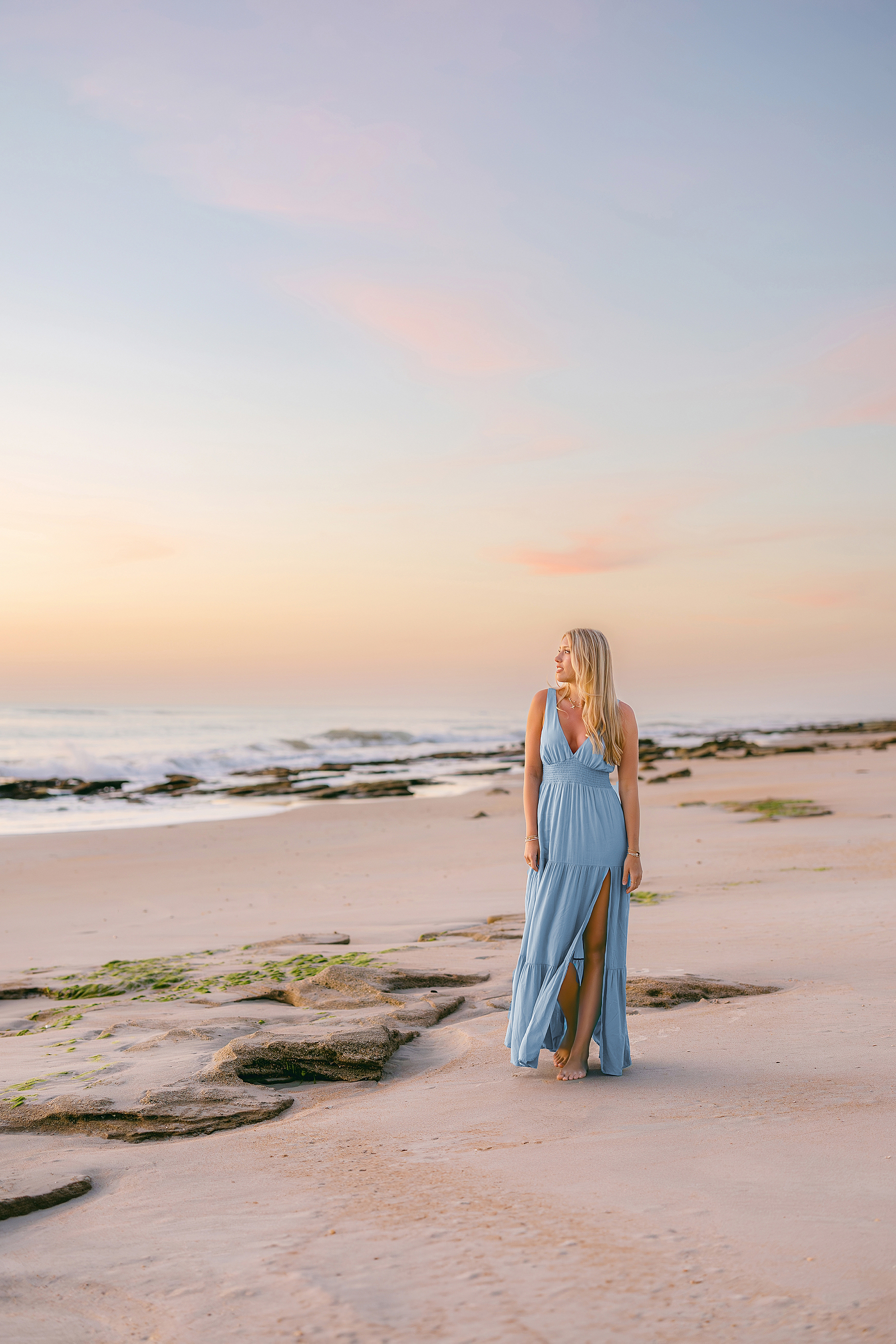 A colorful sunrise portrait of a young grad walking on the beach in a blue dress.