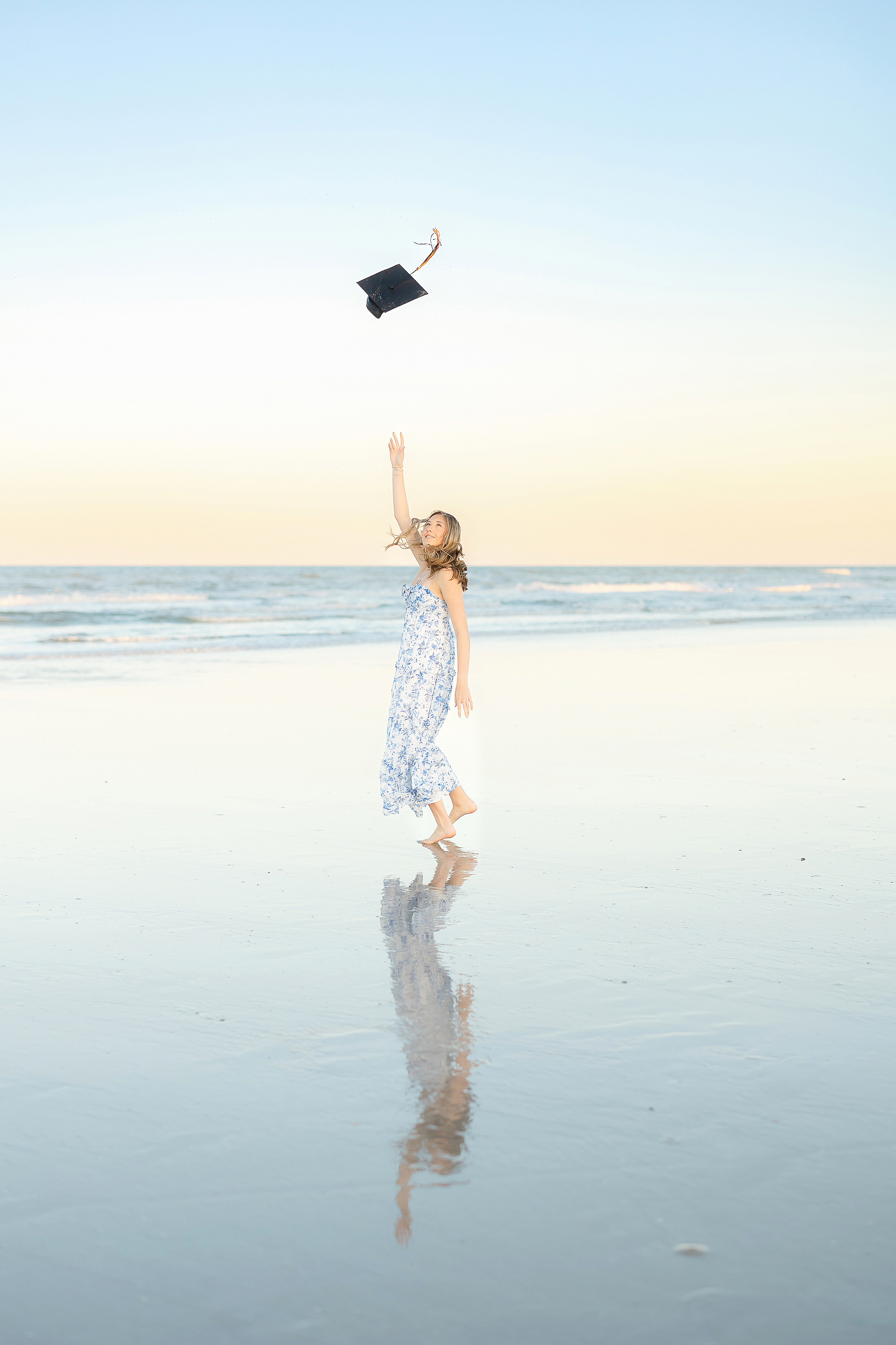 A grad portrait of a young woman in a blue floral dress throwing her cap in the air at sunset on St. Augustine Beach.