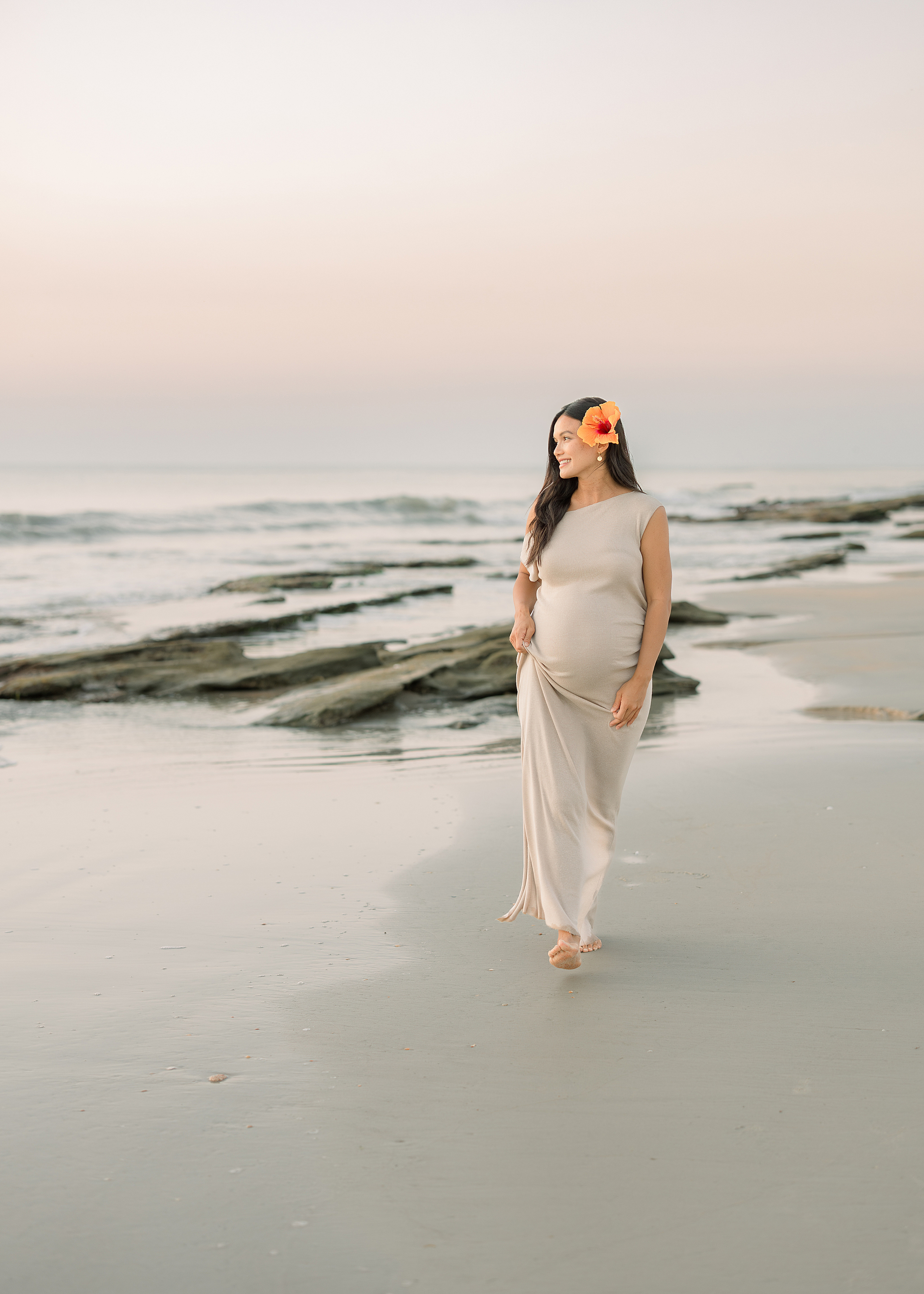 A woman in a cream maxi dress with an orange hibiscus in her hair walks on St. Augustine Beach at sunrise.