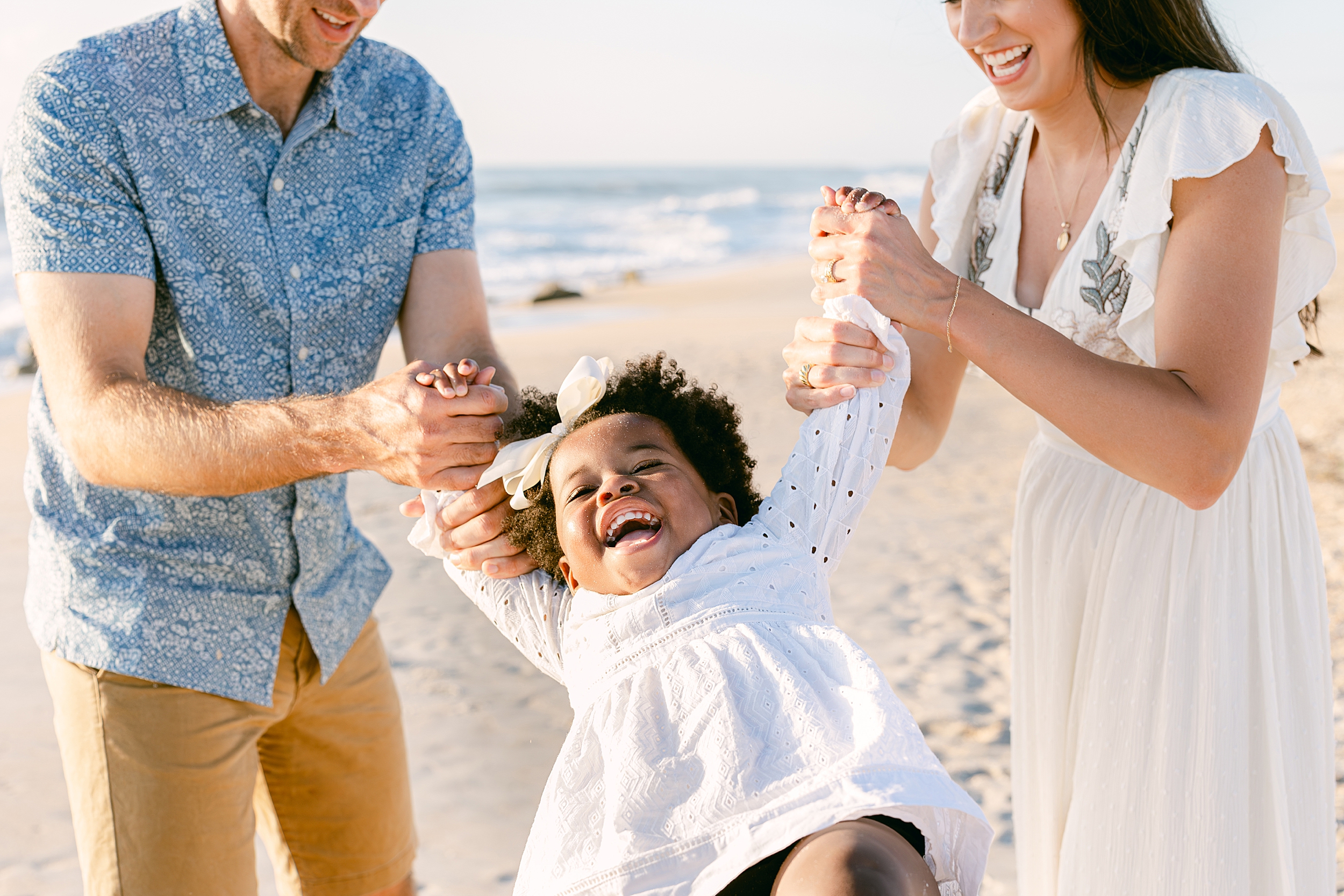 A light and airy beach portrait of a family swinging a little black girl in a white dress.