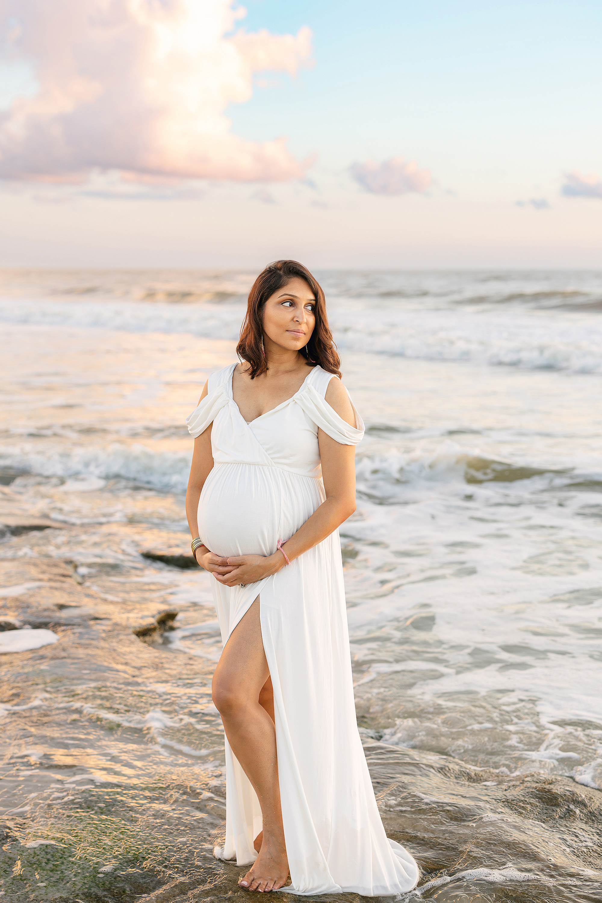 A pregnancy portrait of a woman in a long white dress standing on the rocks at sunrise.