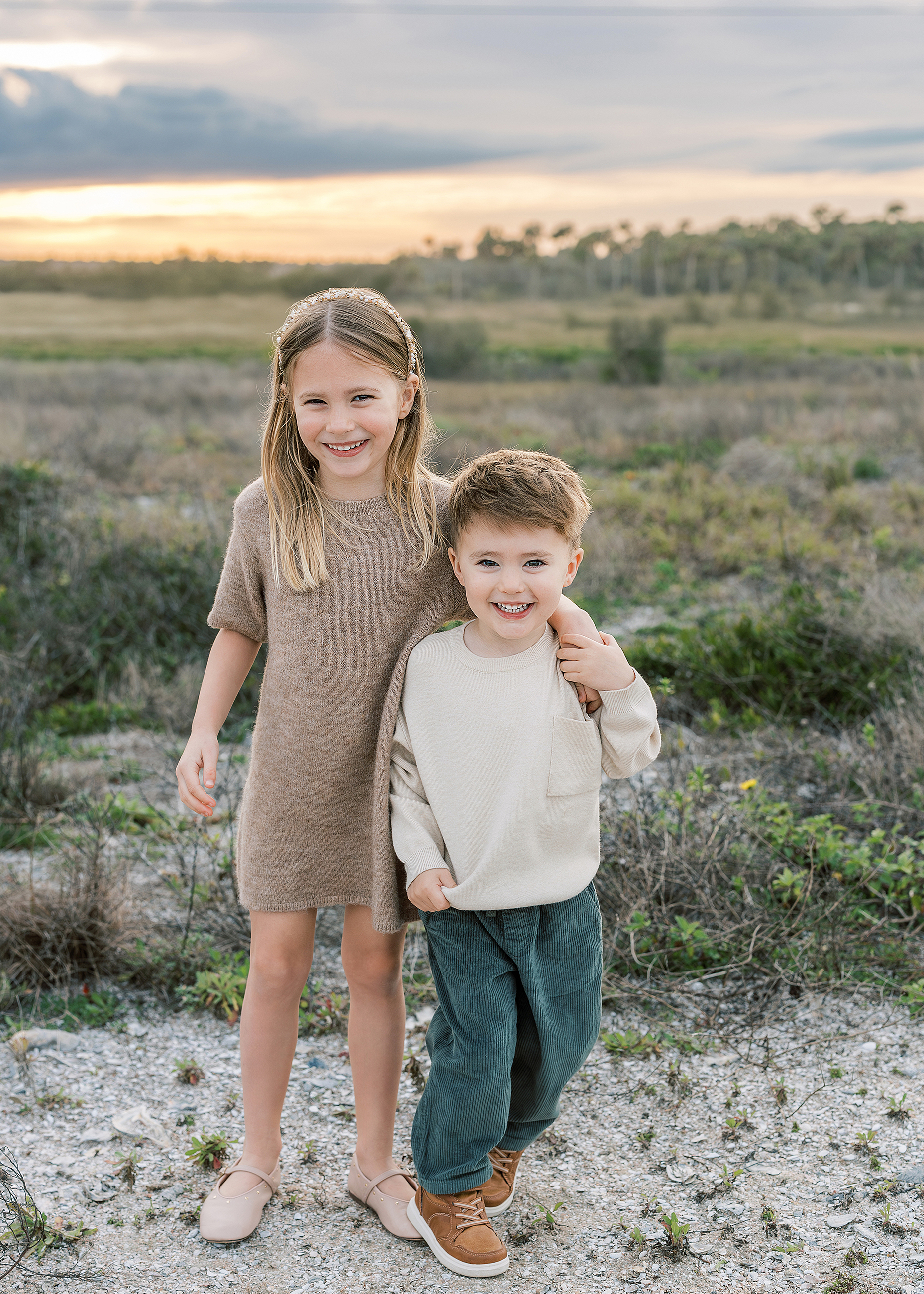 Two little children dressed in green and neutral taupe stand together on the beach.