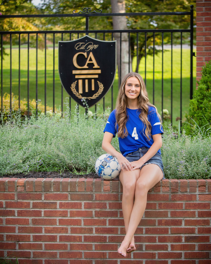 Woman in a blue soccer jersey sitting on a brick wall holding a soccer ball, with a gated sign in the background.