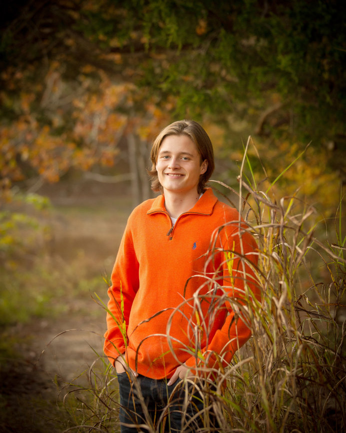 Young person in an orange sweater standing outdoors by tall grass with autumn trees in the background.