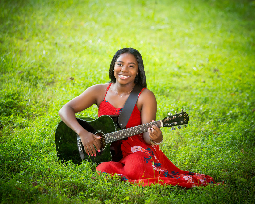 Woman in red dress sitting on grass, playing a black acoustic guitar, smiling.