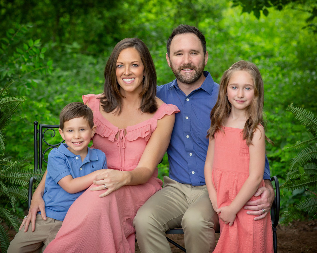 A family of four sits together outdoors, smiling, surrounded by lush greenery.