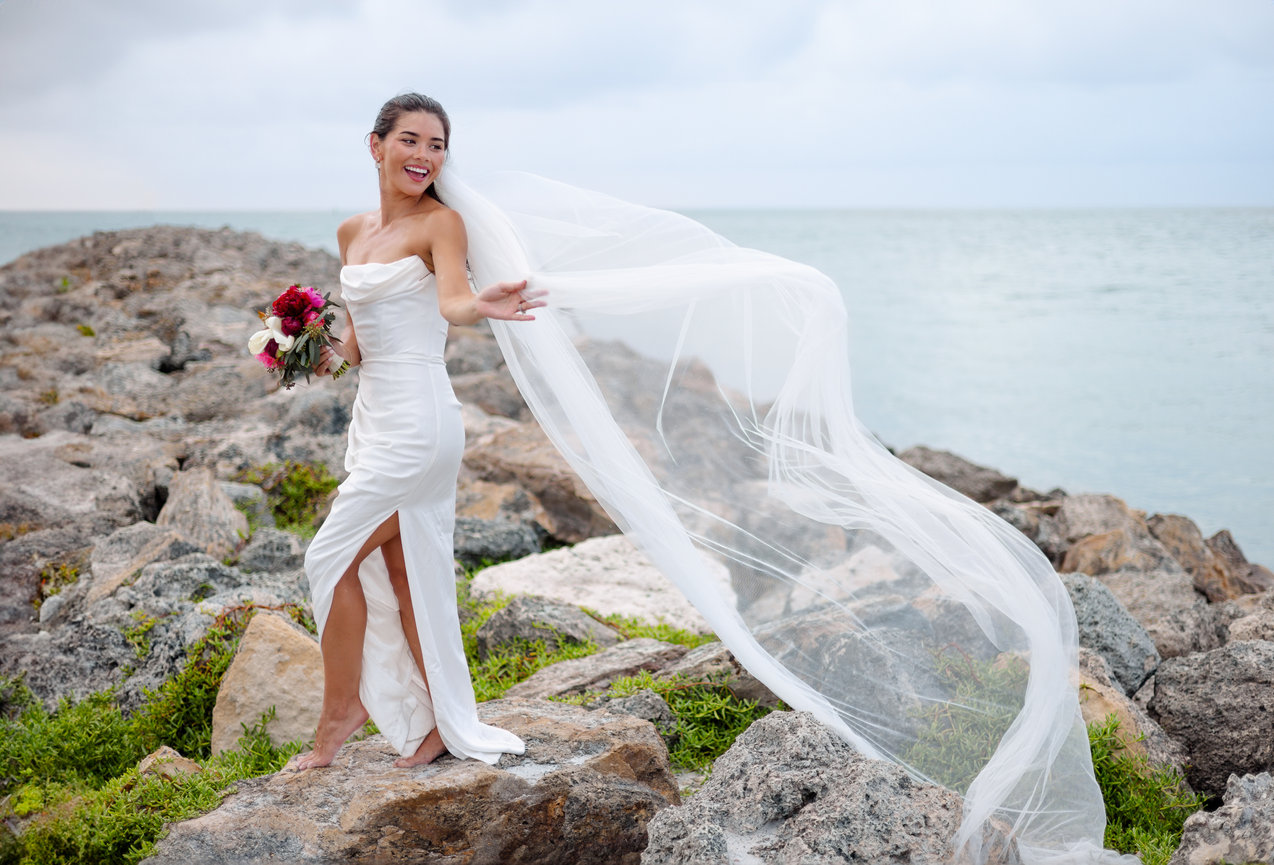 Bride holding bouquet standing on rocks at Sand Key Beach with veil blowing in the breeze