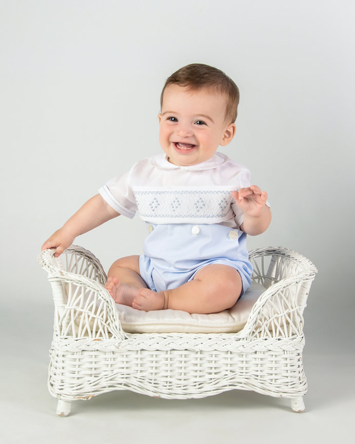 Smiling baby sitting in a white wicker chair wearing a light blue outfit.