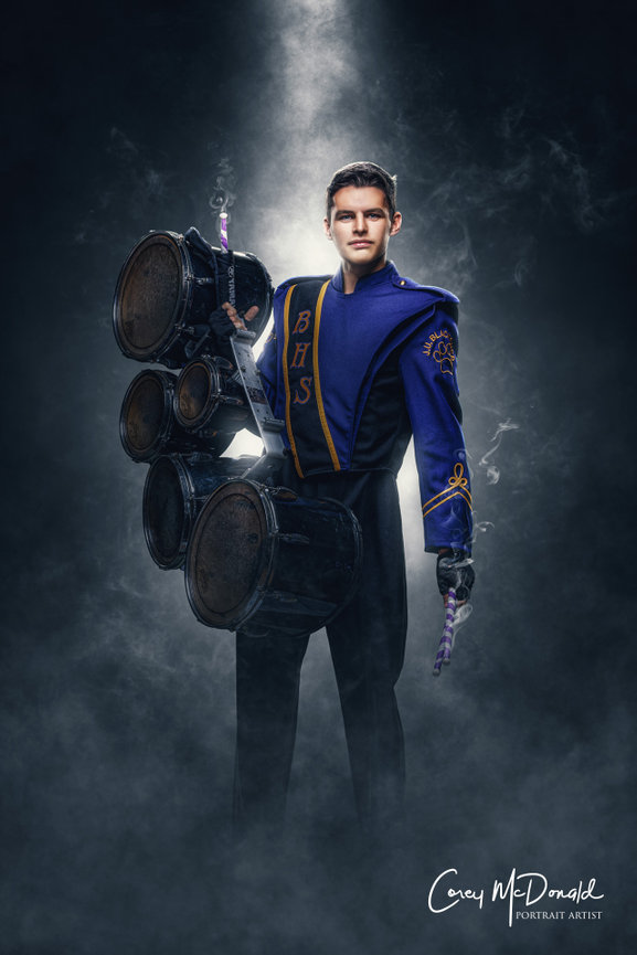 Marching band member in blue and black uniform holds multiple drums in a spotlight, with smoke swirling around.