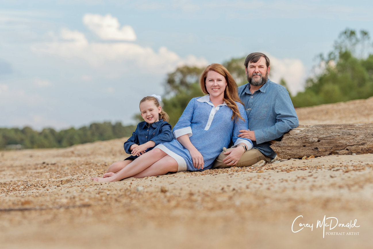 A family of three sits on a sandy beach, smiling, with trees and clouds in the background.