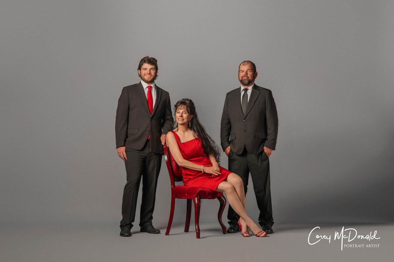 A woman in a red dress sits on a chair, flanked by two men in suits, against a neutral backdrop.