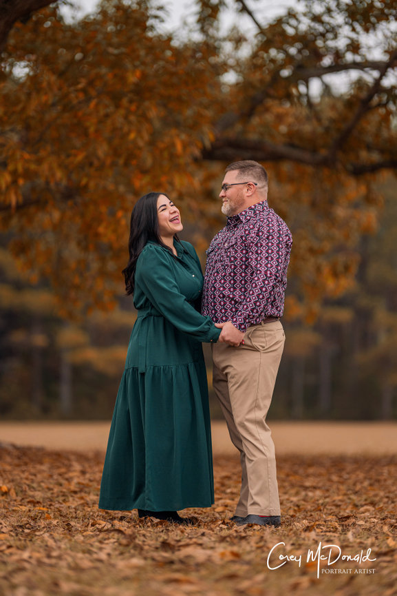 A couple laughing together under a large tree with autumn leaves.