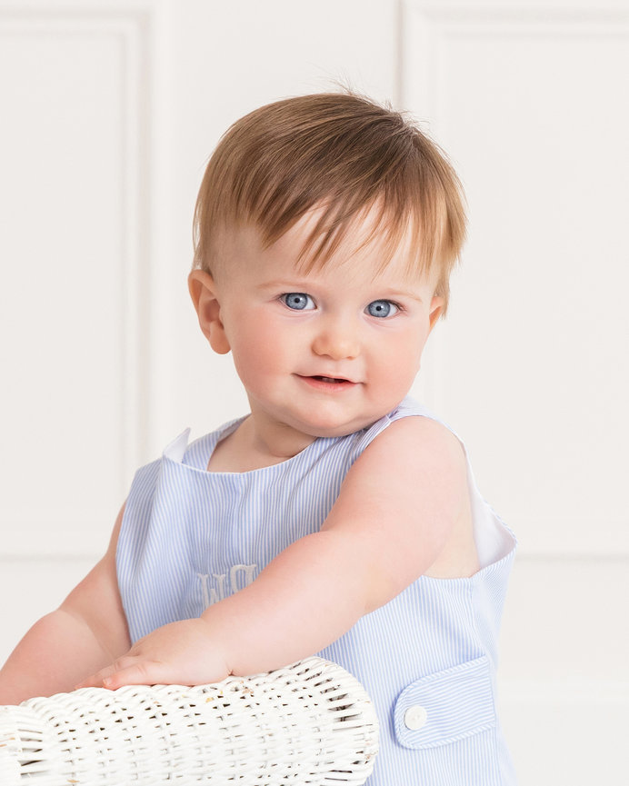 Smiling baby in a light blue outfit standing near a white wicker chair indoors.
