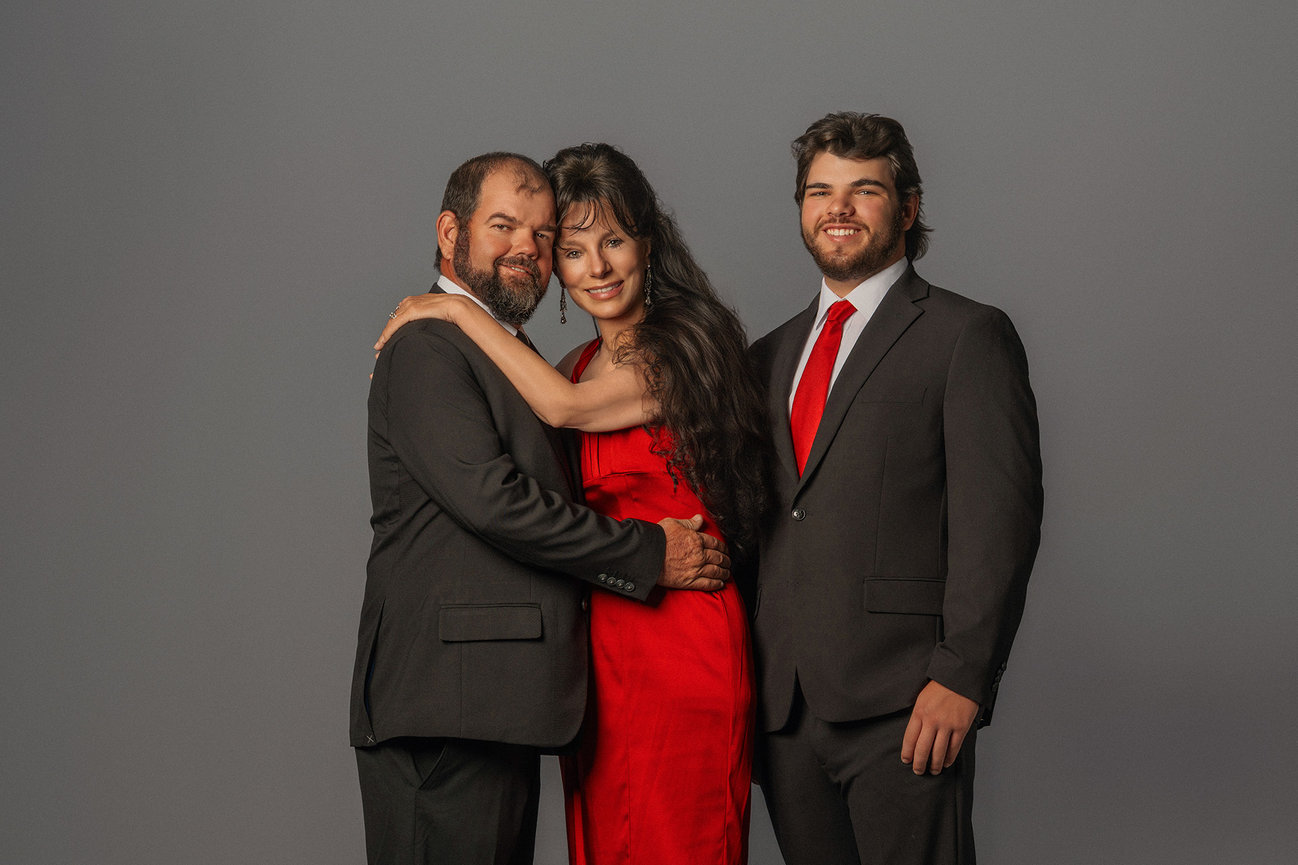 Three people posing; two men in dark suits and a woman in a red dress, smiling against a gray background.