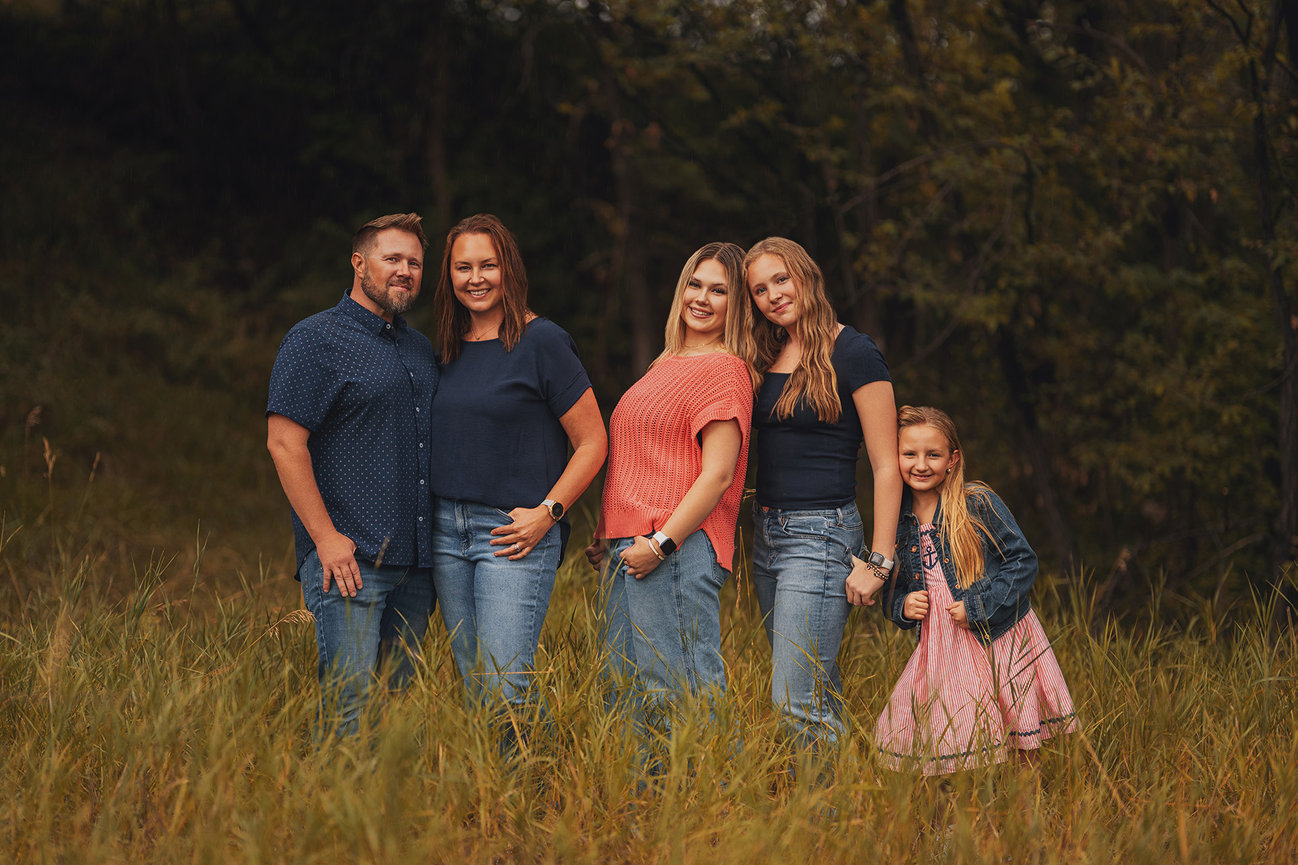 A family of five poses together outdoors in a grassy area with trees in the background.