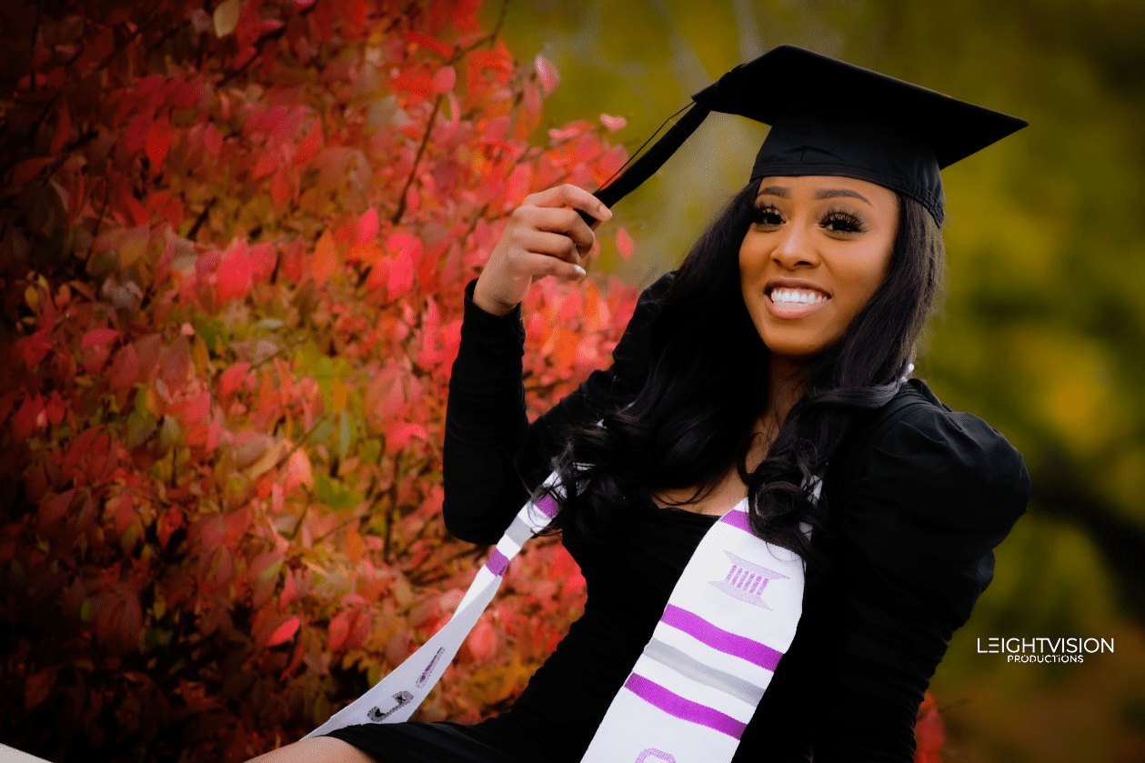 A woman in a graduation gown smiles proudly while posing for a photo, celebrating her academic achievement.