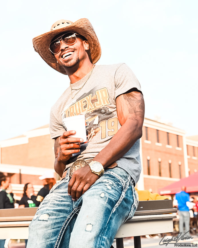 A cheerful man in a cowboy hat and jeans, smiling warmly at the camera.