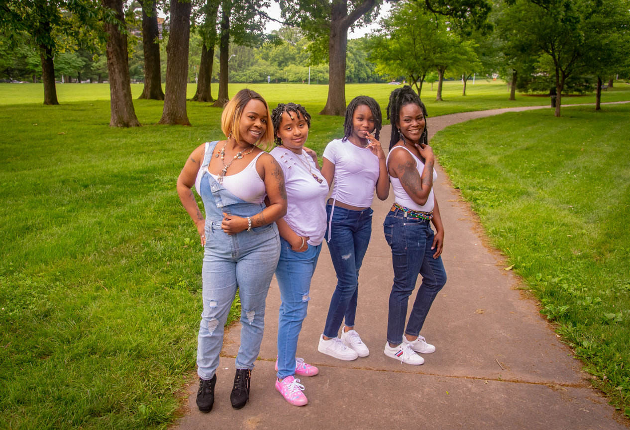 Four women posing together on a pathway in a green park, wearing casual outfits and smiling.