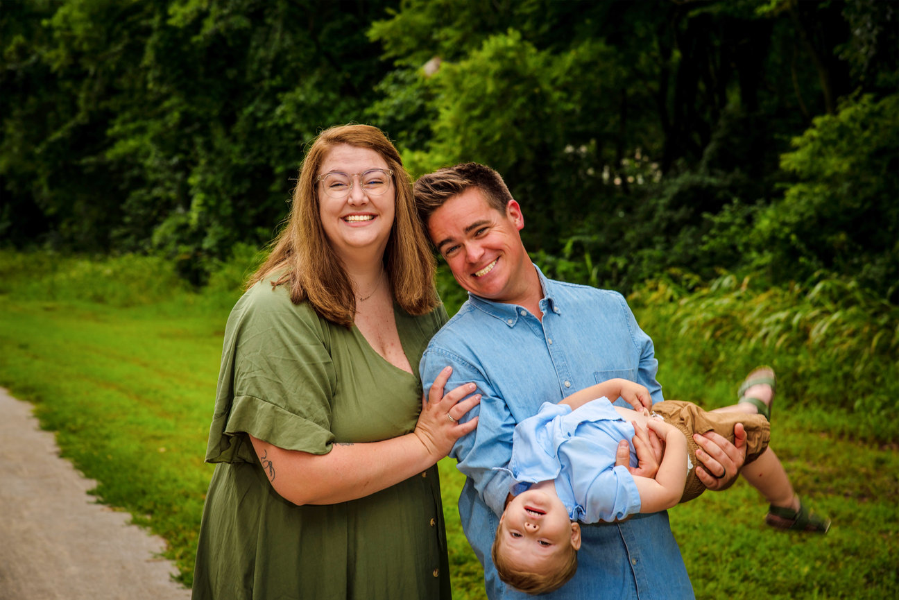 Happy family portrait with parents playfully holding their smiling child outdoors in a lush green setting.