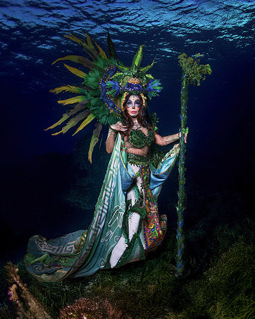 Woman in ornate headdress and flowing costume underwater with fish swimming above