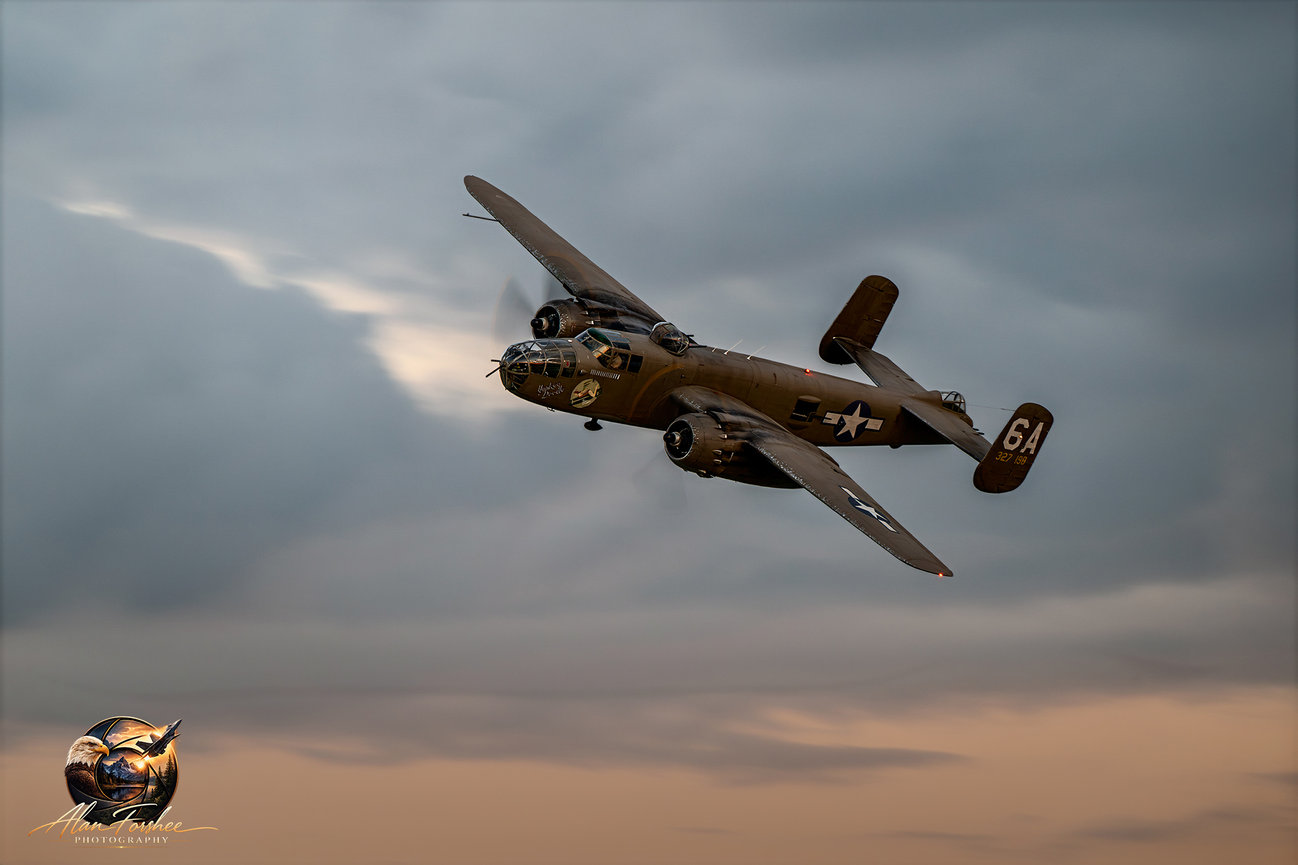 Historic B-25 Mitchell bomber Yankee Doodle banking in flight against a dramatic gray and warm-toned evening sky.