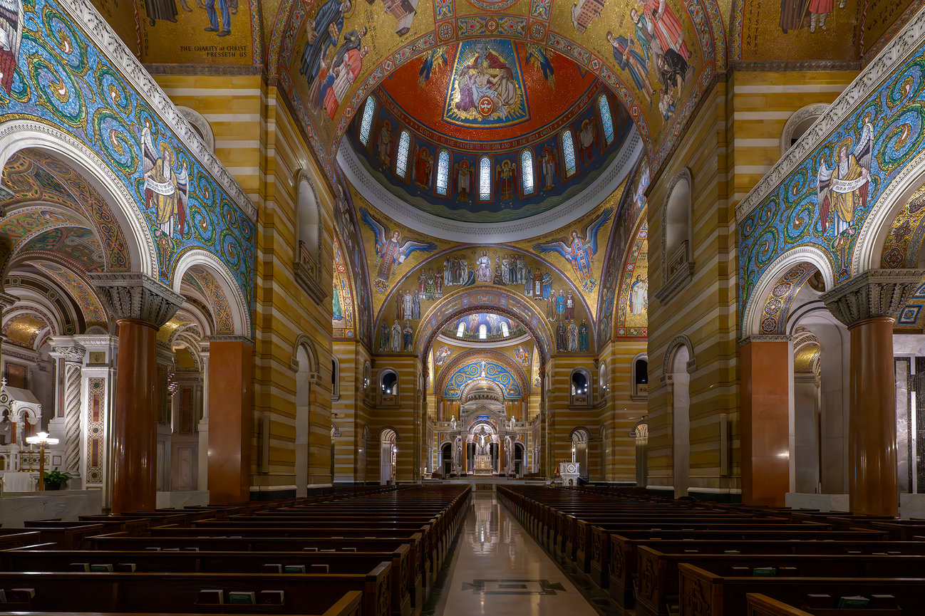 Wide interior view of the Cathedral Basilica of Saint Louis showing colorful mosaics, ornate arches, wooden pews, and the central aisle leading to the sanctuary.