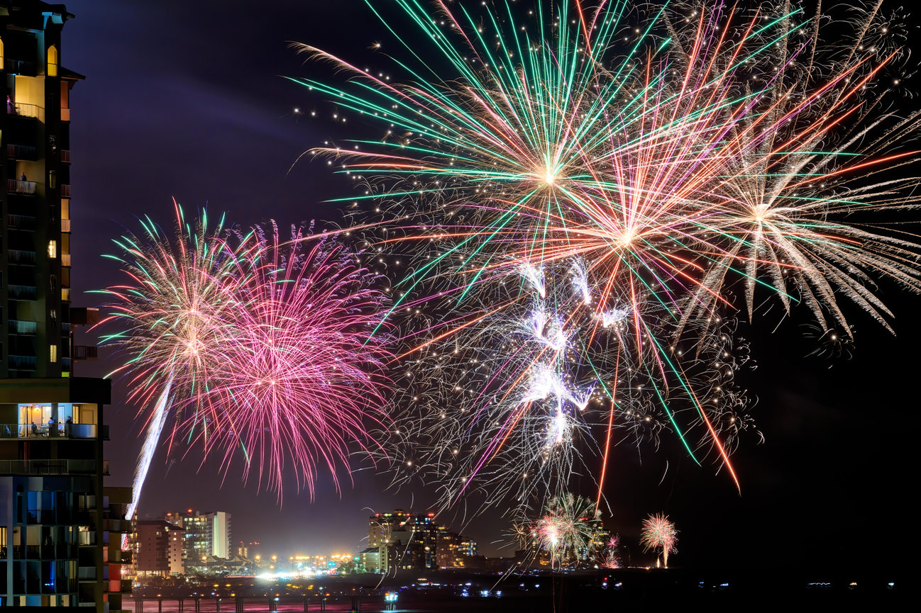 Colorful fireworks burst over a city skyline at night beside a high-rise building.