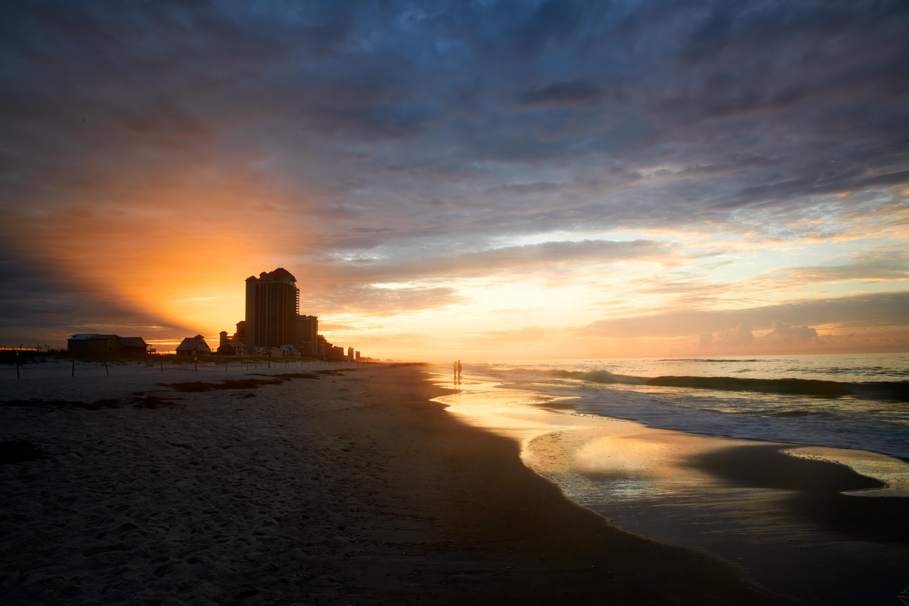 Sunset over a quiet beach with ocean waves, a distant skyline, and two people near the shore