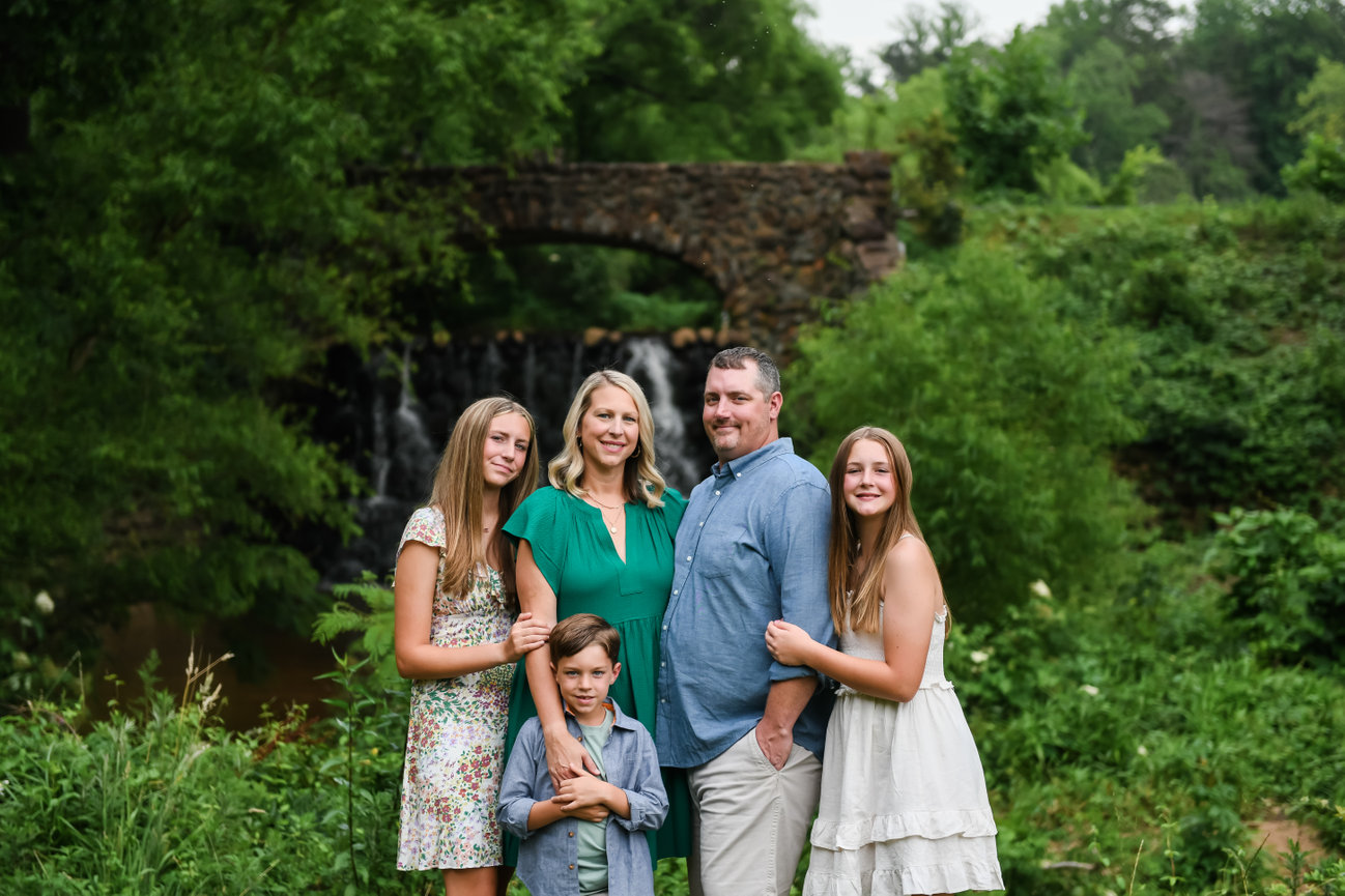 Family of five standing together in front of a stone bridge and waterfall surrounded by lush greenery during a summer photo session at Reynolda Gardens in Winston-Salem, North Carolina