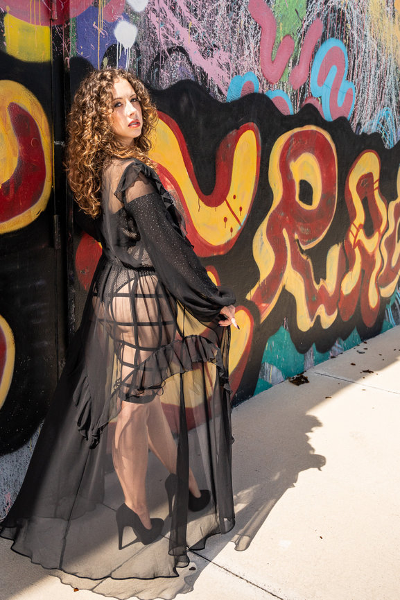 Person in a sheer black outfit poses confidently, standing beside a vibrant graffiti wall.