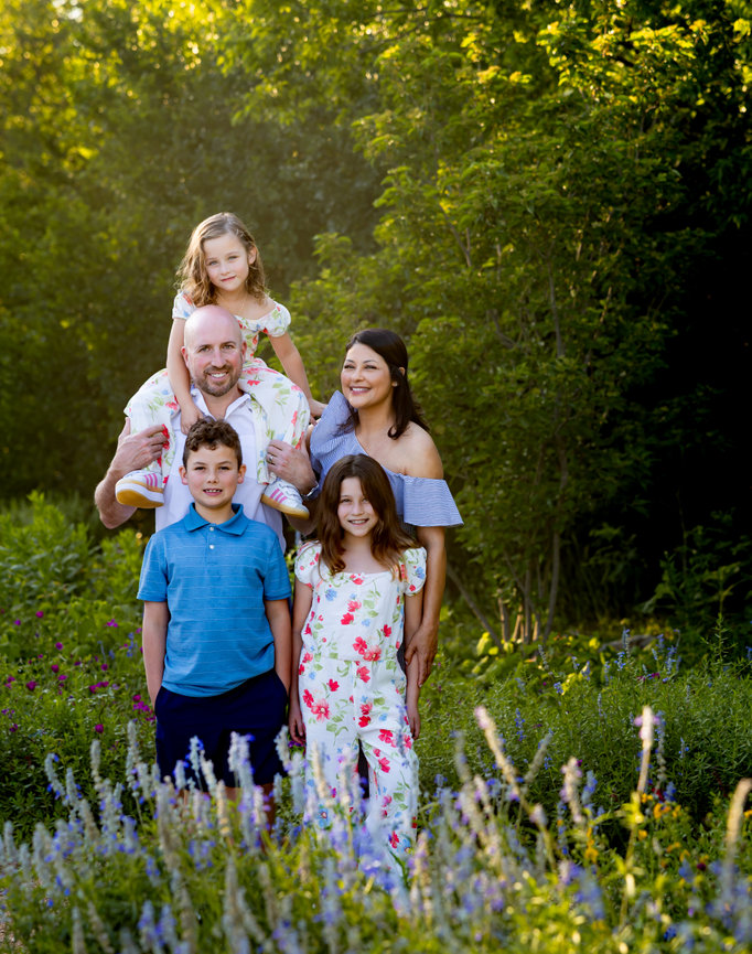 Family of five posing in a sunny garden with wildflowers and lush green trees