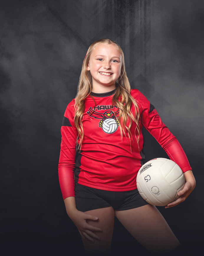 Young girl in a red volleyball jersey holding a white volleyball, smiling against a dark background.