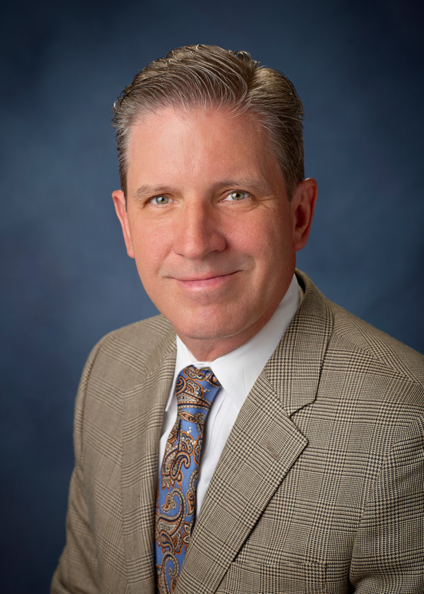 Professional headshot of a man in a suit and paisley tie against a blue background