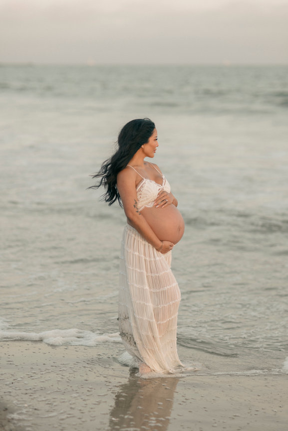 orange county Pregnant woman in a flowing white dress standing on the beach, waves gently lapping at her feet.