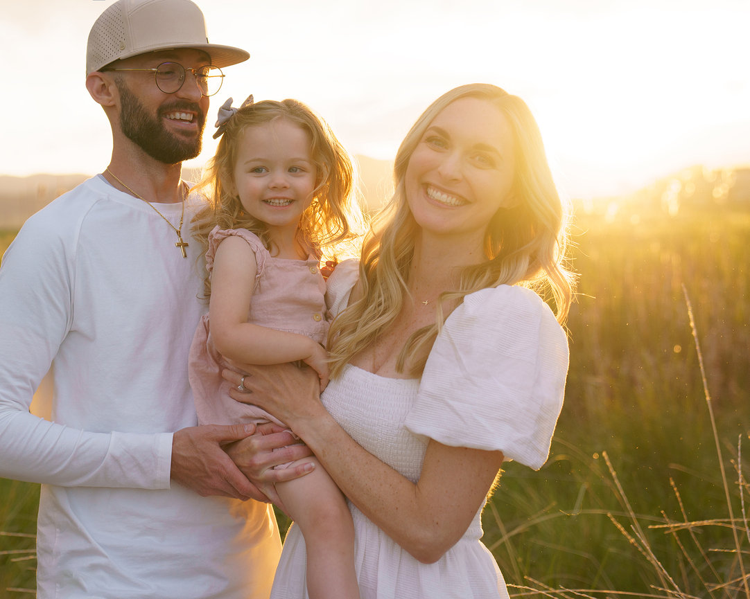 Smiling family of three in a sunny field, the woman holding a young child, with a warm sunset in the background.
