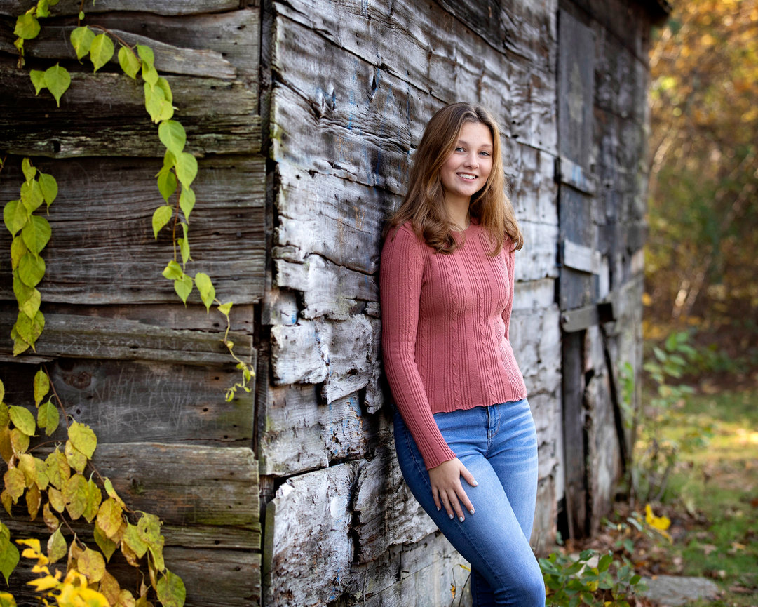 Senior in a red sweater and jeans leans against a rustic wooden shed, surrounded by autumn foliage.