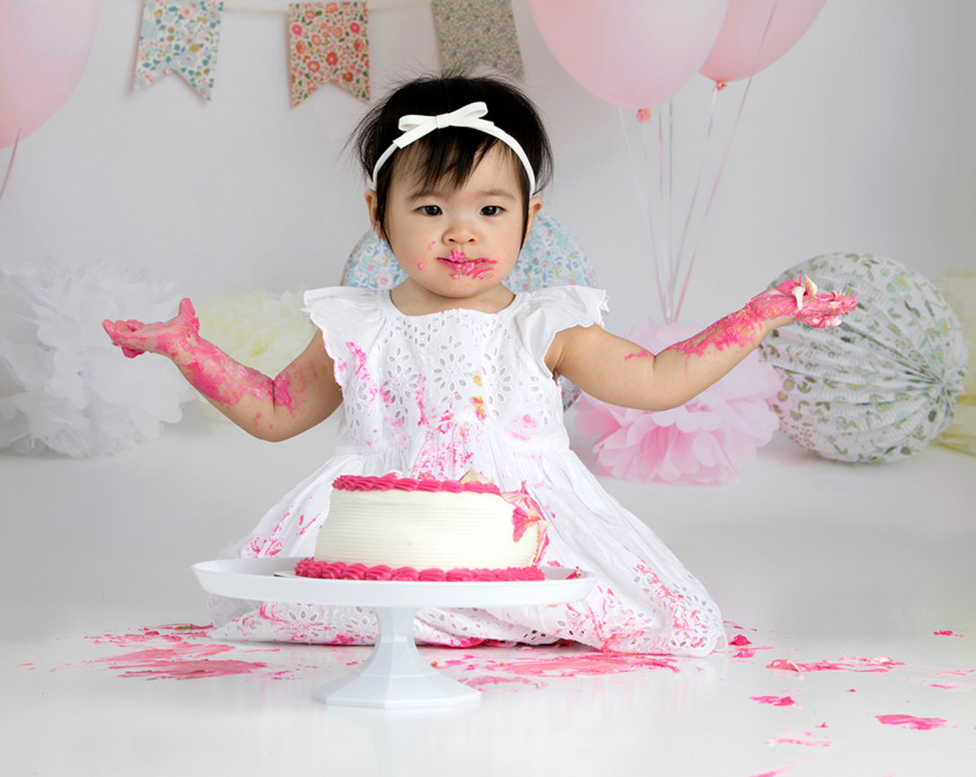 Baby in a white dress with pink frosting on her face and arms sits behind a cake on a stand in a festive setting.