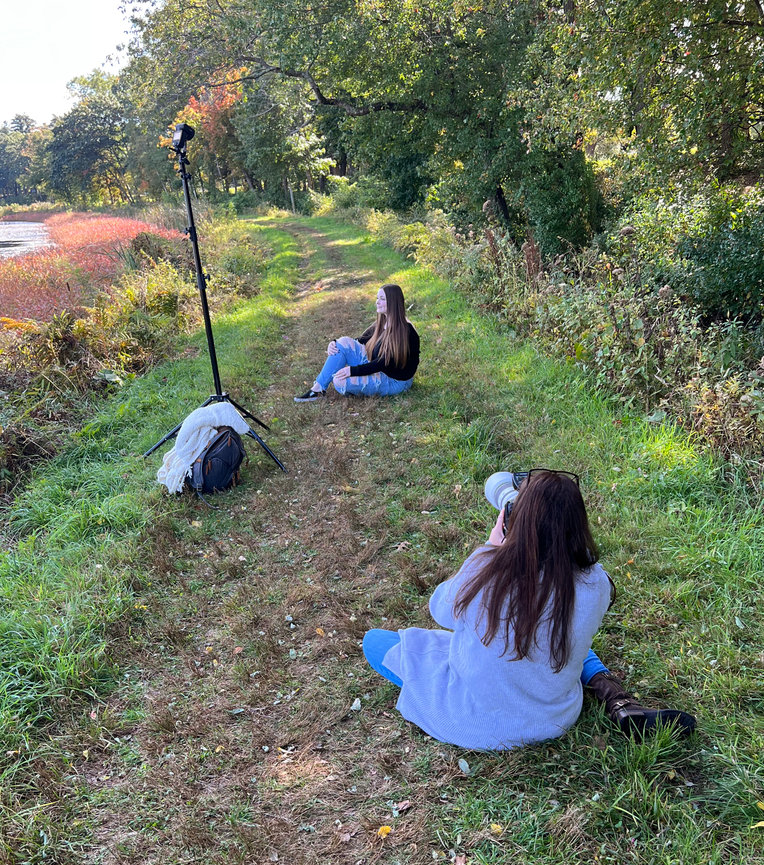 Paula Swift photographing a high school senior sitting on a grassy path near a lake, with trees in the background at Farm Pond.