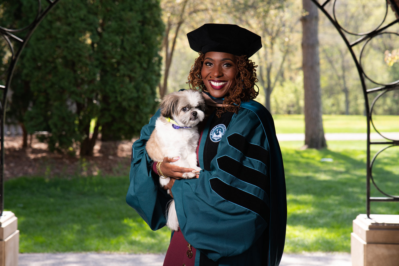 Smiling graduate in cap and gown holding a small dog outdoors under a garden arch