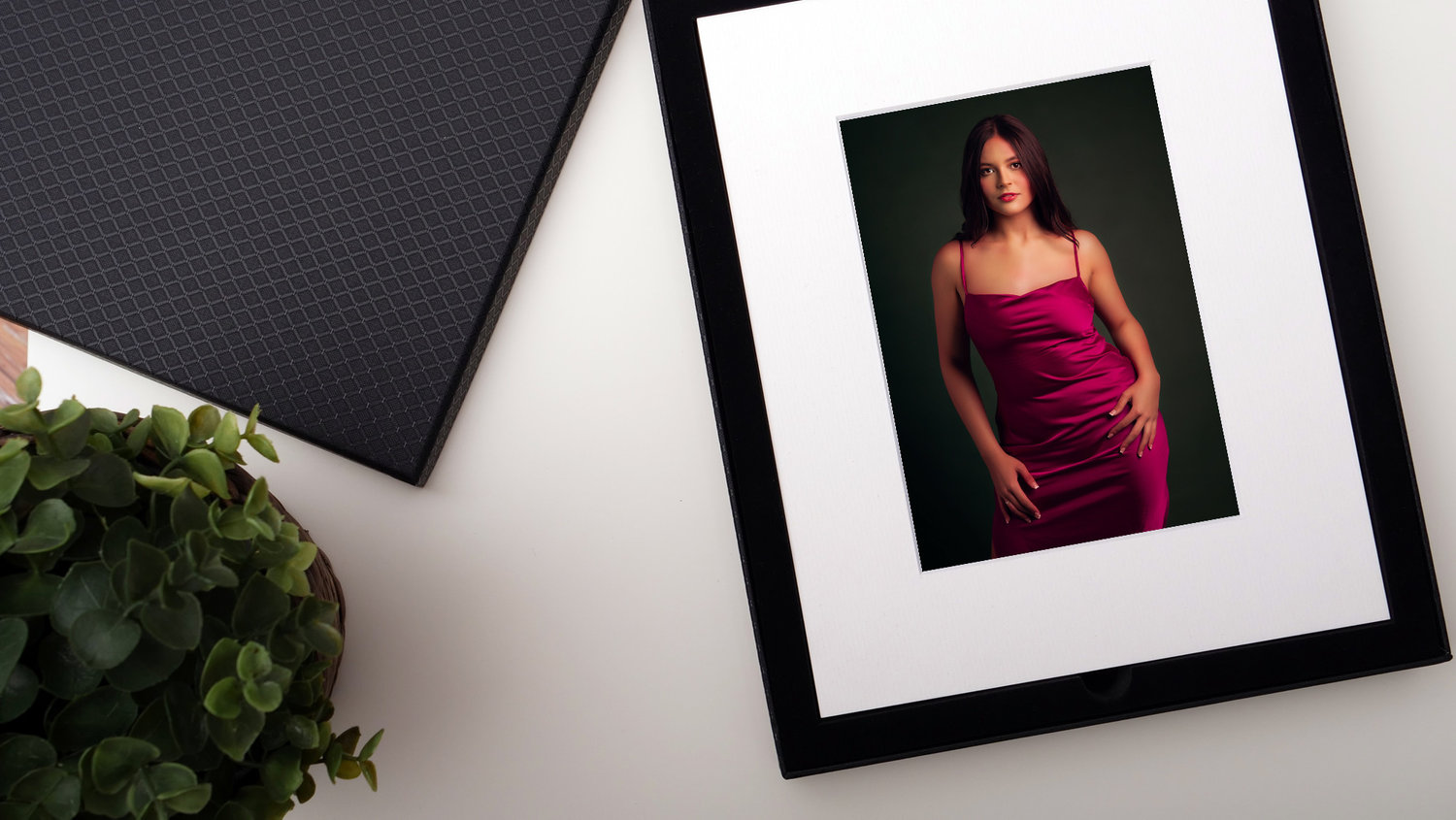 Framed photo of a woman in a pink dress on a white surface, next to a plant and a textured dark pad.
