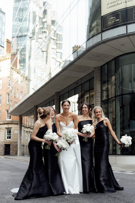 Modern bridal party portrait in Manchester; bridesmaids in chic black dresses holding white floral bouquets in an editorial street setting.