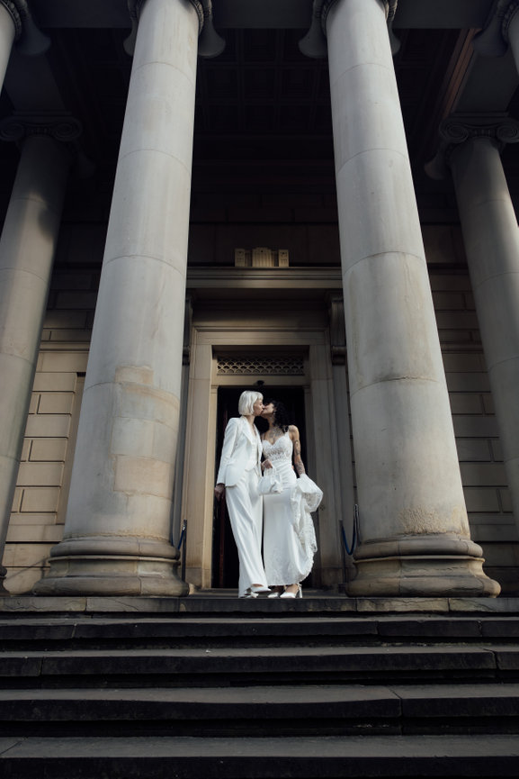Editorial wedding photography of two brides kissing on the stone steps, framed by the grand neoclassical columns of Manchester Art Gallery.
