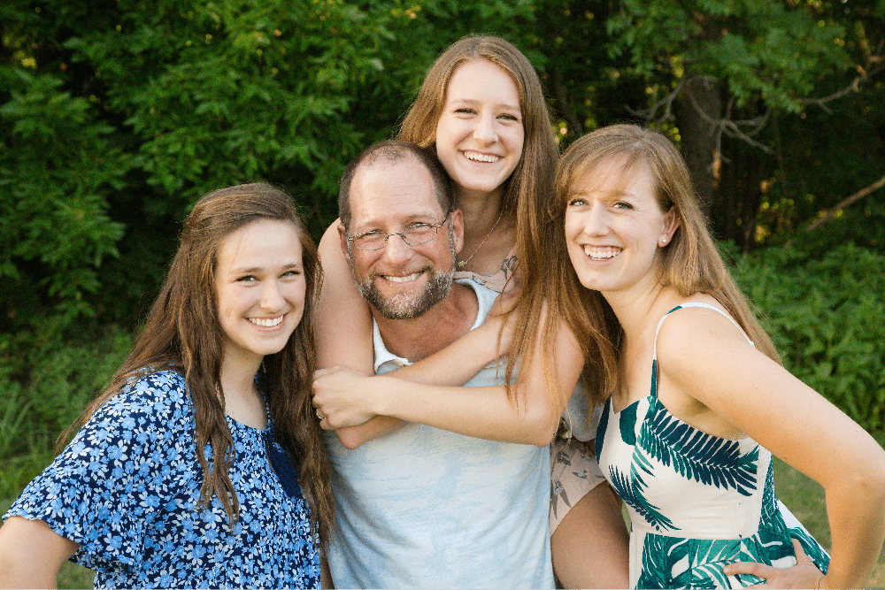 Smiling family of four posing outdoors in front of green foliage during a family photo session in Blue Springs, MO photographed by Case Studios Photography