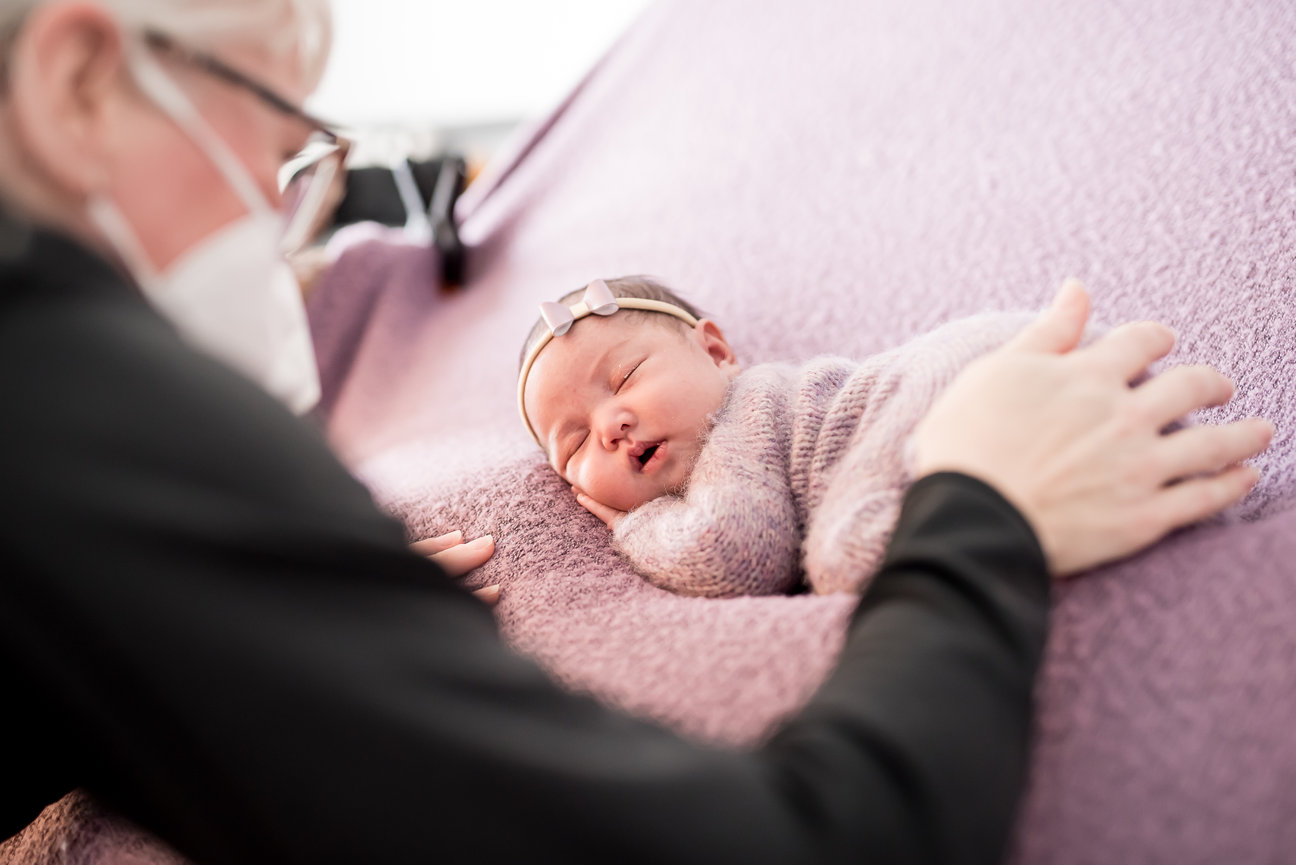 Person gently cradling a sleeping newborn wrapped in a purple blanket, resting on a matching backdrop.