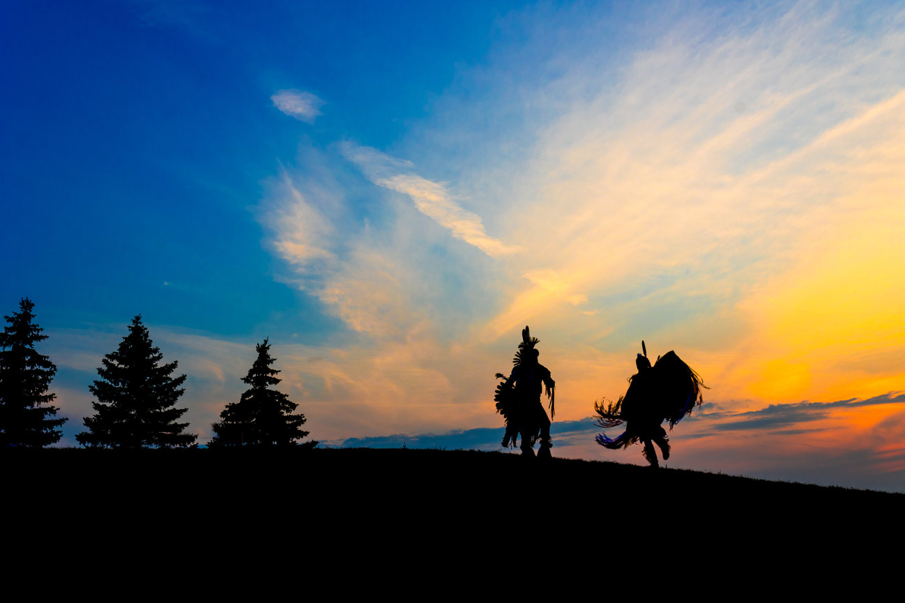 Two silhouetted dancers in traditional attire perform against a vibrant sunset, with trees outlined in the foreground.