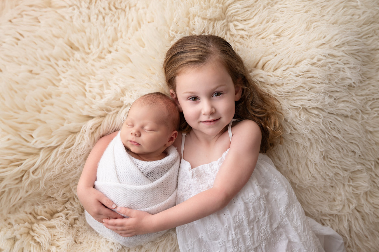 Toddler sister lovingly cuddling swaddled baby on soft, textured rug in professional newborn portrait