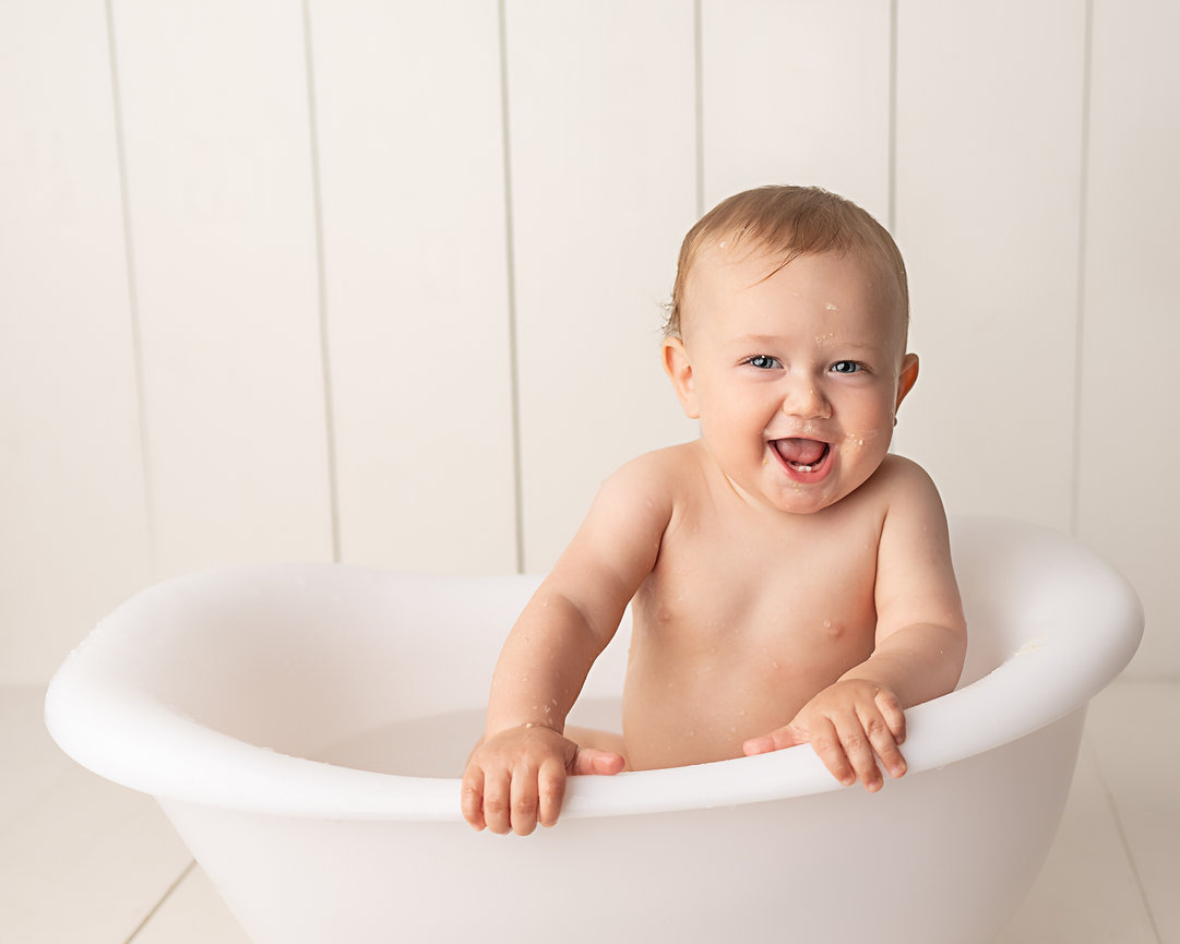 Baby laughing while rinsing off frosting in white bathtub after Wall Twp photoshoot