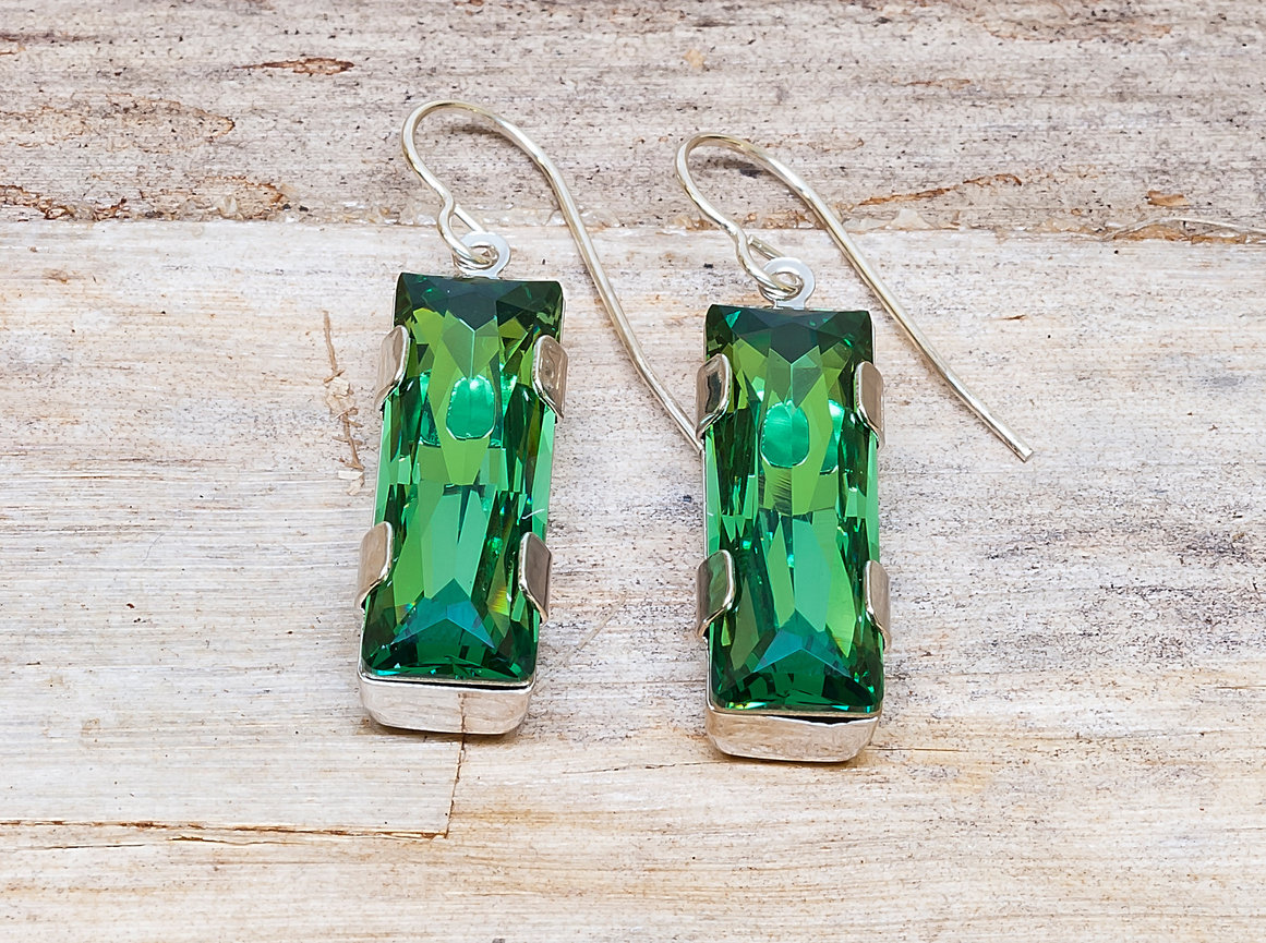 Pair of silver hook earrings featuring rectangular, faceted green gemstones in prong settings, displayed on a rustic wooden surface