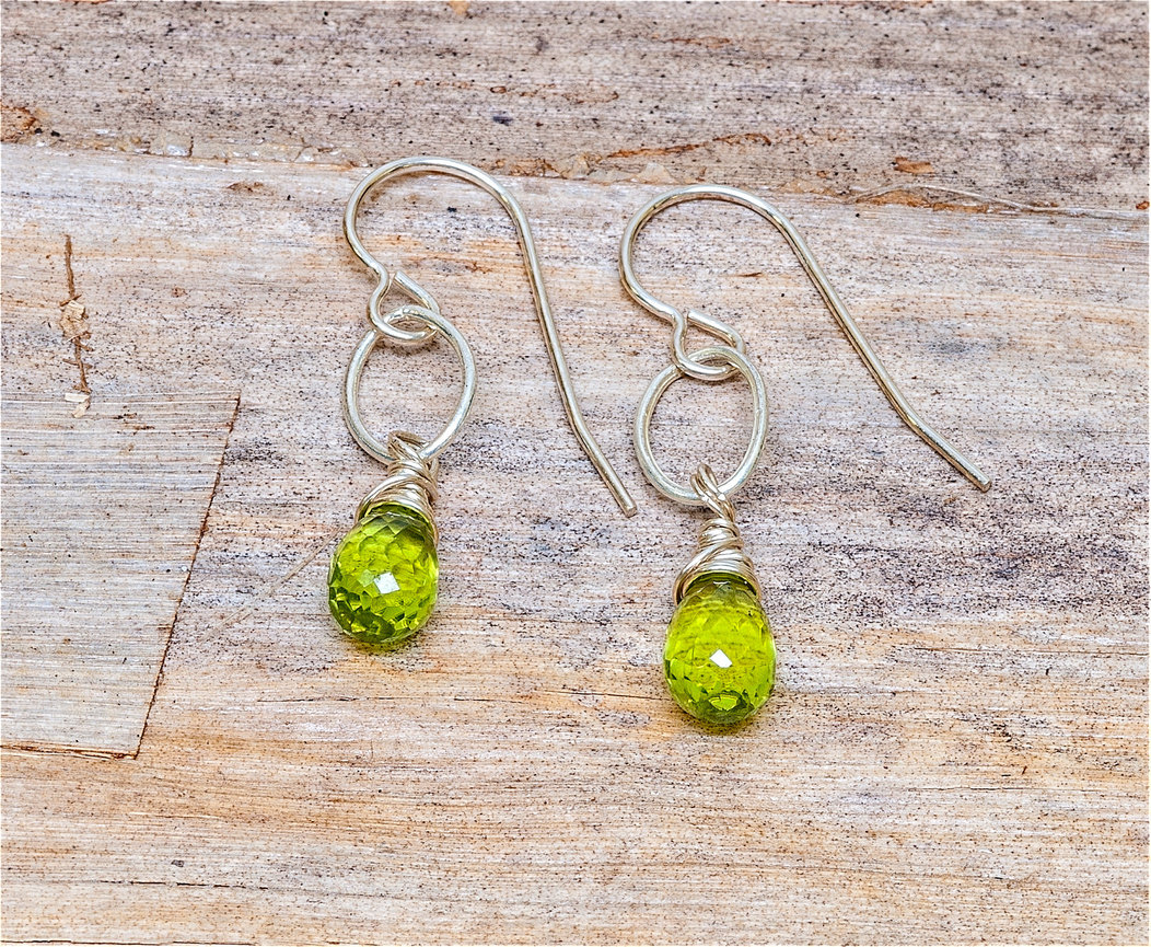 Pair of handmade dangle earrings featuring faceted teardrop-shaped peridot gemstones wrapped in silver wire, suspended from oval silver links and hook ear wires, displayed on a rustic wooden surface