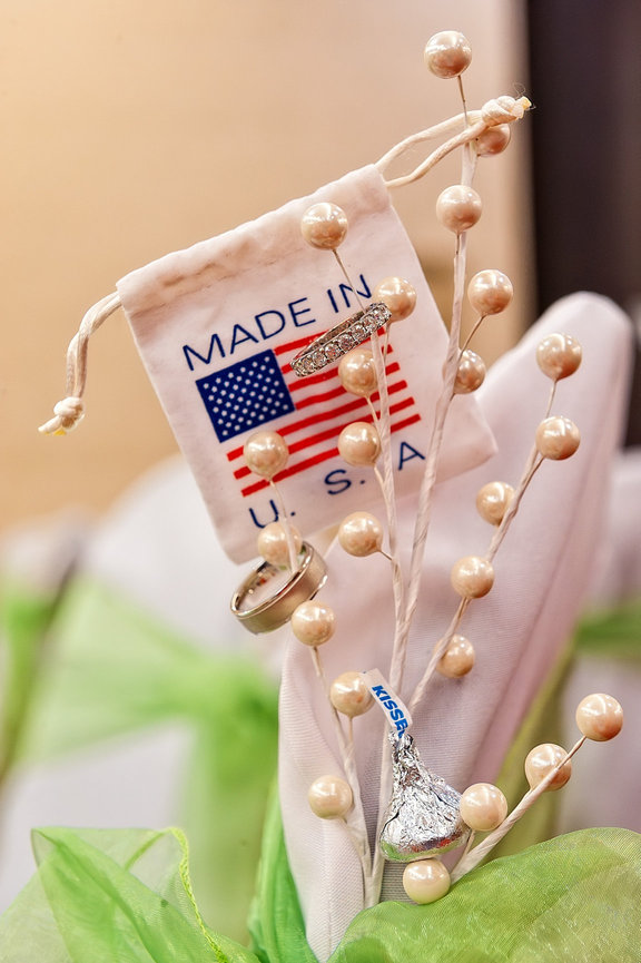 Wedding rings displayed on pearl-decorated stems alongside a small “Made in U.S.A.” drawstring bag and a Hershey’s Kiss, with green and white fabric in the background.